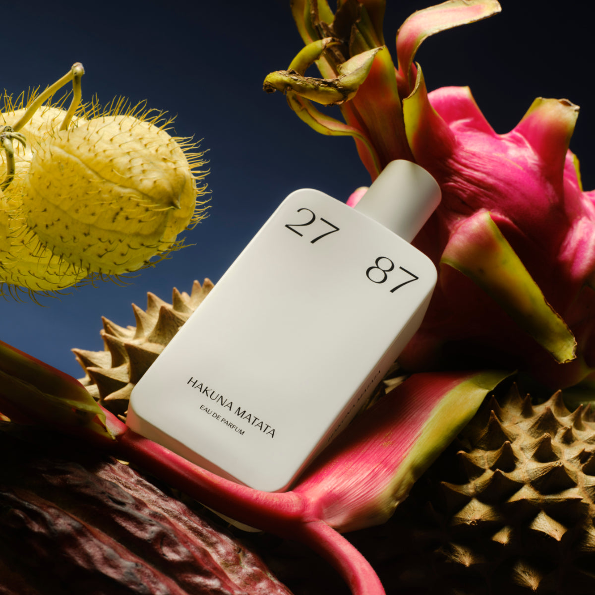 27 87 Perfumes - Hakuna Matata Flakon mit minimalistischer Gestaltung vor exotischen Blüten und Früchten auf dunklem Hintergrund