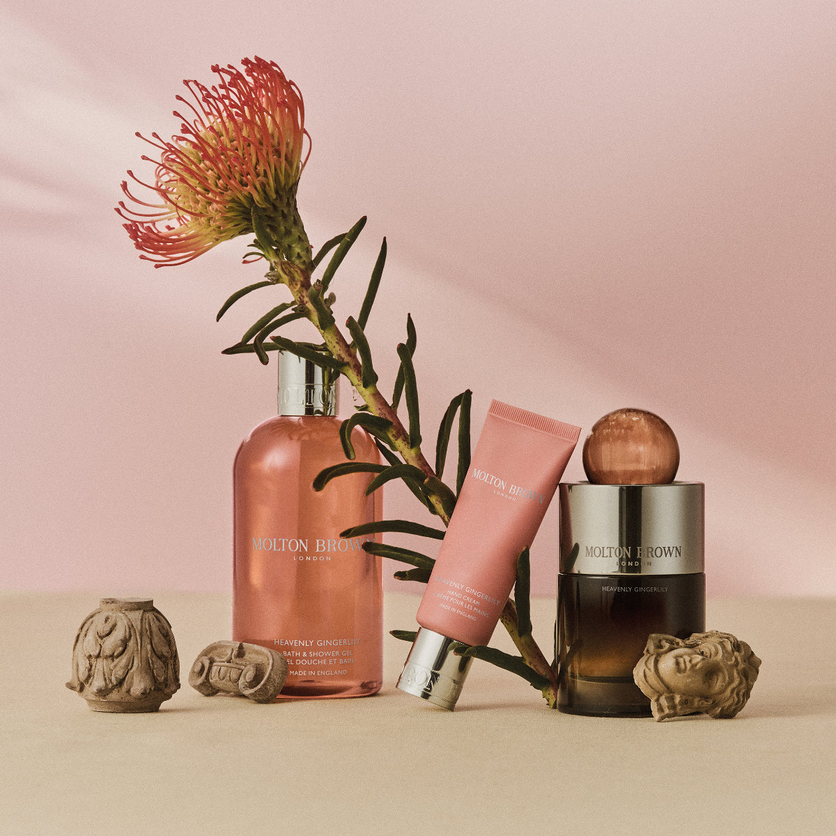 Molton Brown - Heavenly Gingerlily Hand Cream in rosa Tube, arrangiert mit Blüte und Duftflakons vor zartem Hintergrund