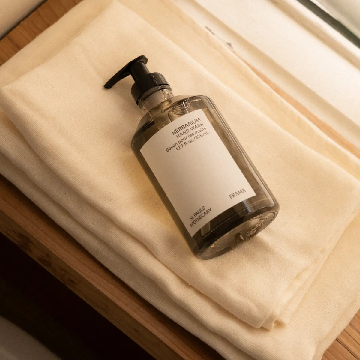 FRAMA - Herbarium - Hand Wash milde Flüssigseife in klarem Spender mit schwarzem Pumpspender auf beige Stoff gelegt