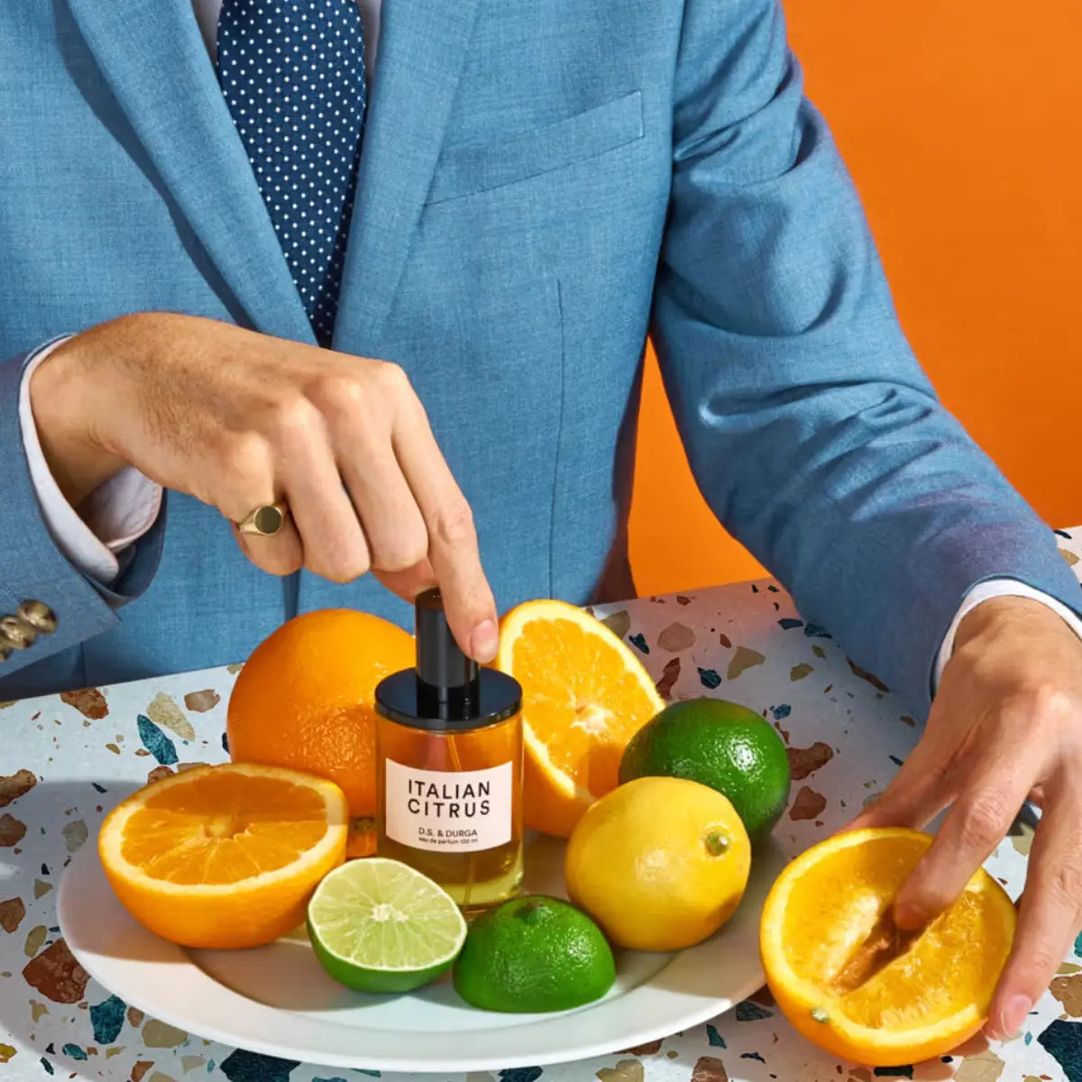 D.S. & Durga - Italian Citrus Parfümflakon auf Teller mit Zitrusfrüchten vor orangem Hintergrund und blauem Anzug