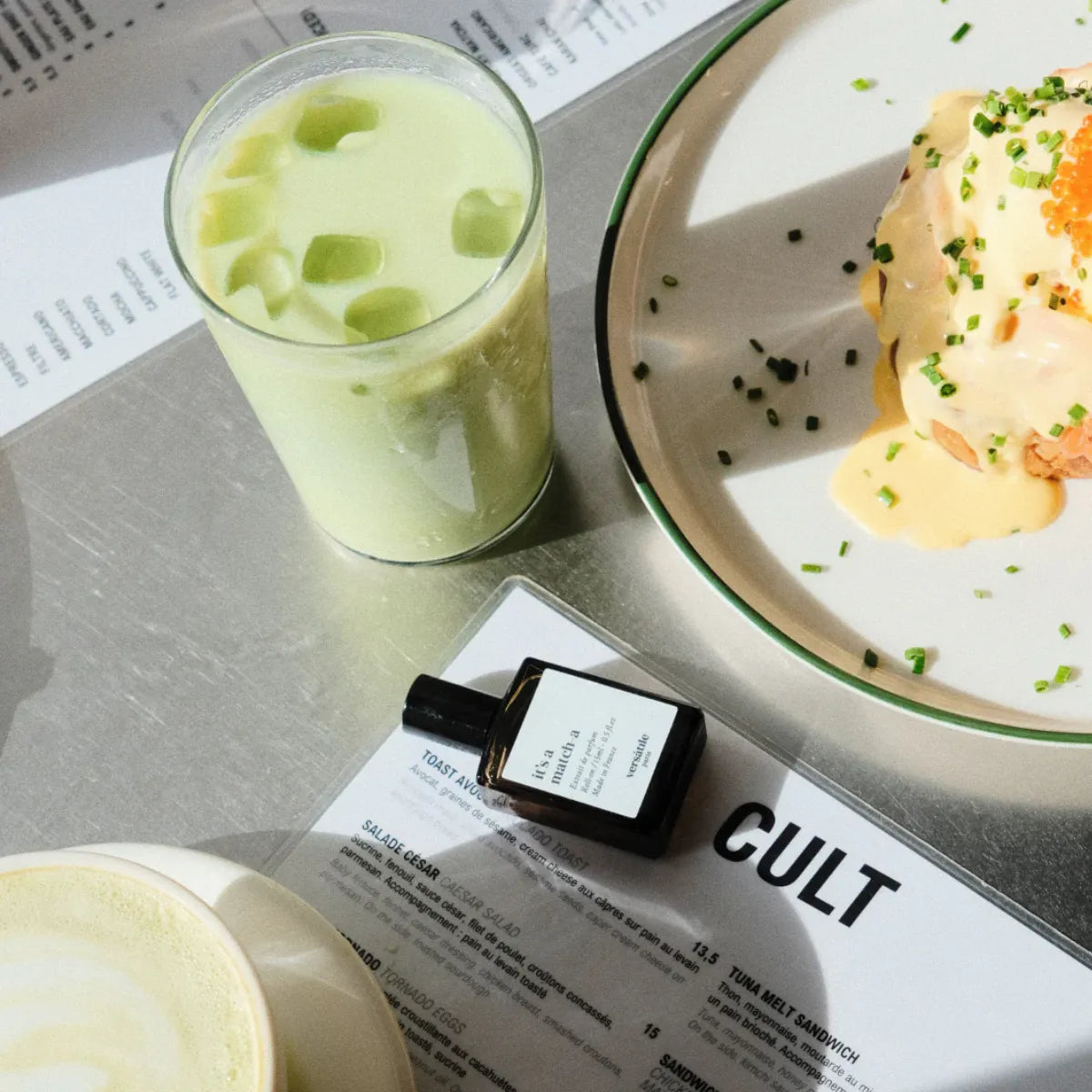 Versatile - It’s a Match-A Parfümflakon neben grünem Matcha-Getränk und Teller mit brunchgericht auf hellem Untergrund