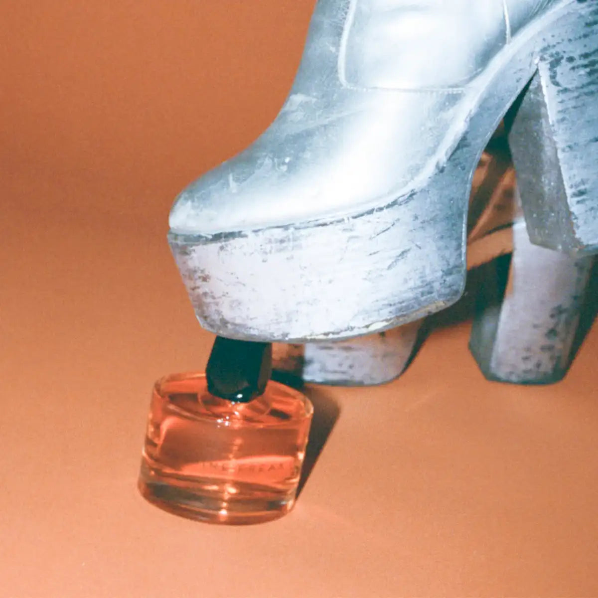 Room 1015 - Jasmine Freak Parfümflakon liegt auf orangem Boden neben auffälligem silbernem Plateau-Schuh