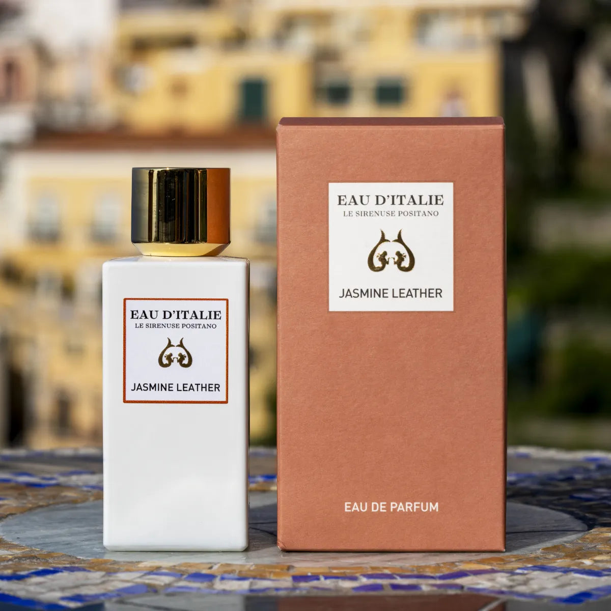 Eau d'Italie - Jasmine Leather weißer Flakon mit goldenem Verschluss neben brauner Verpackung vor unscharfem Hintergrund