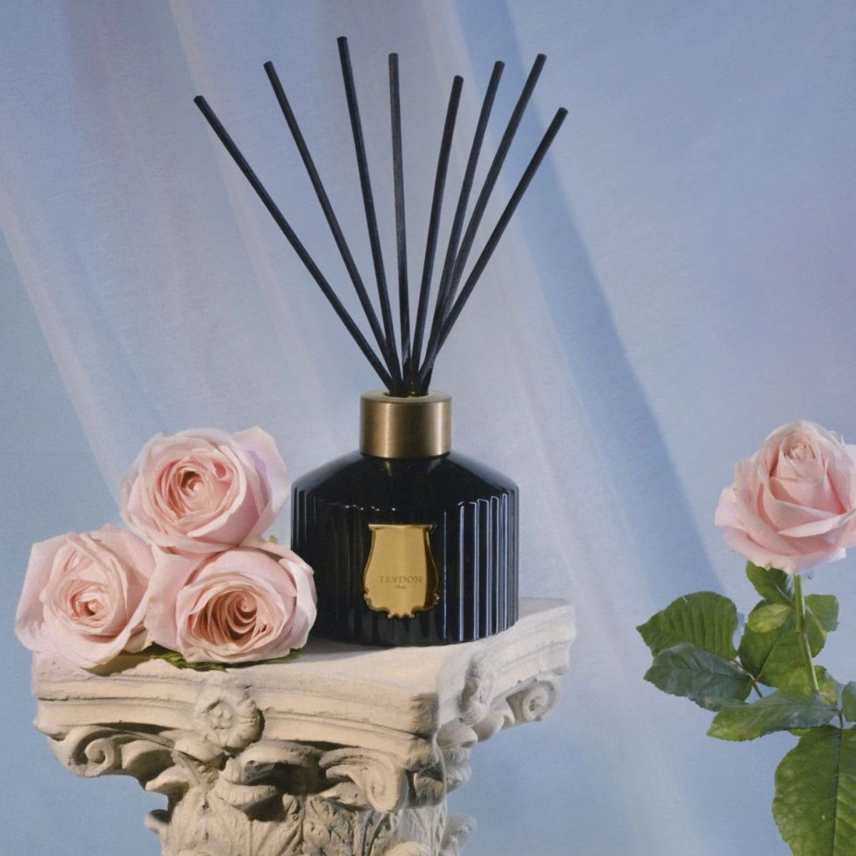 Trudon - Reed Diffusor - Joséphine mit schwarzem geriffeltem Flakon und goldfarbenem Verschluss auf dekorativem weißen Sockel mit Rosen