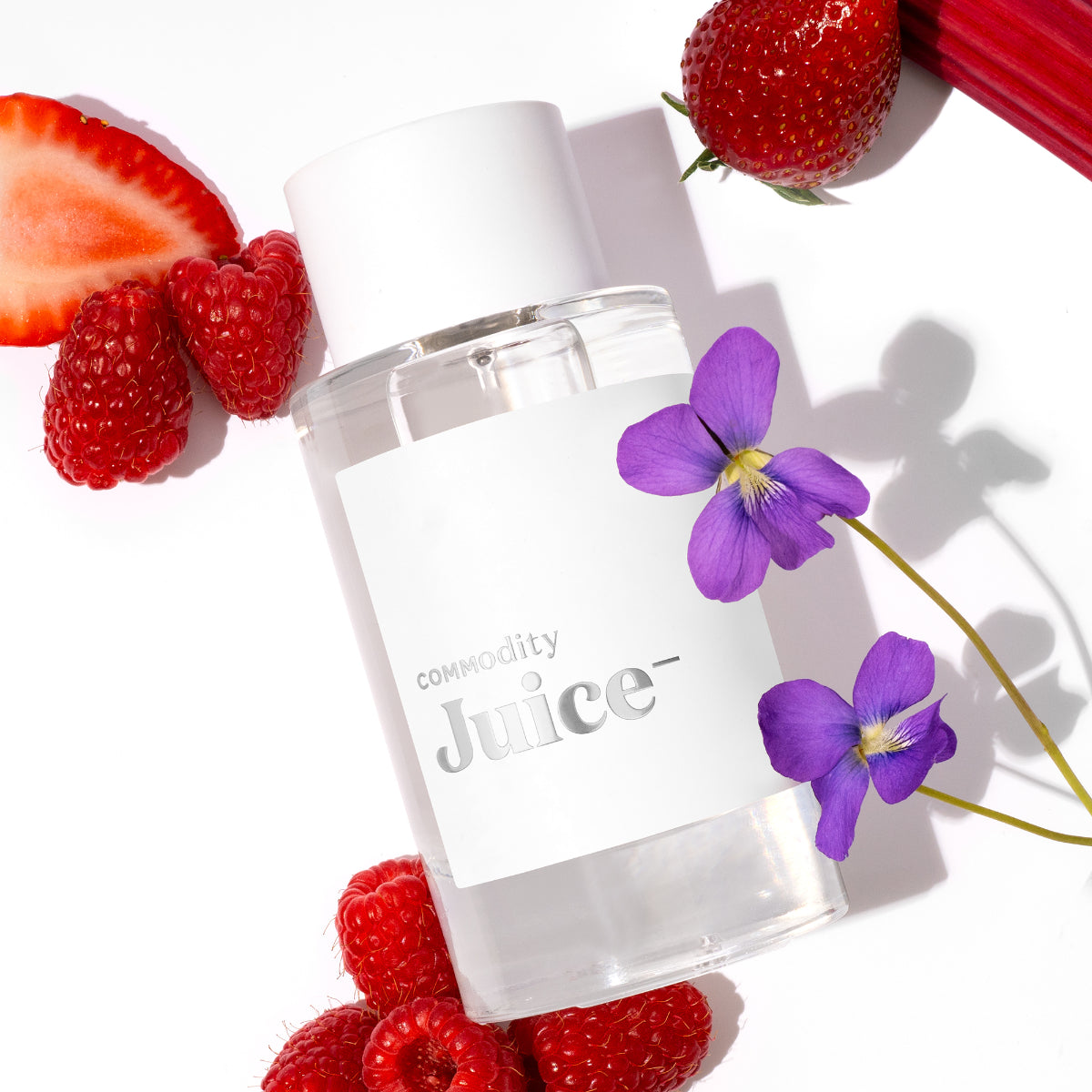 Commodity - Juice- klarer Flakon mit weißem Deckel und Etikett umgeben von Erdbeeren, Himbeeren, Rhabarber und Veilchenblüten