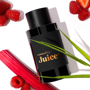 Commodity - Juice+ schwarzer Flakon mit kupferfarbenem Logo umgeben von Rhabarber, Erdbeeren und Himbeeren auf weißem Hintergrund