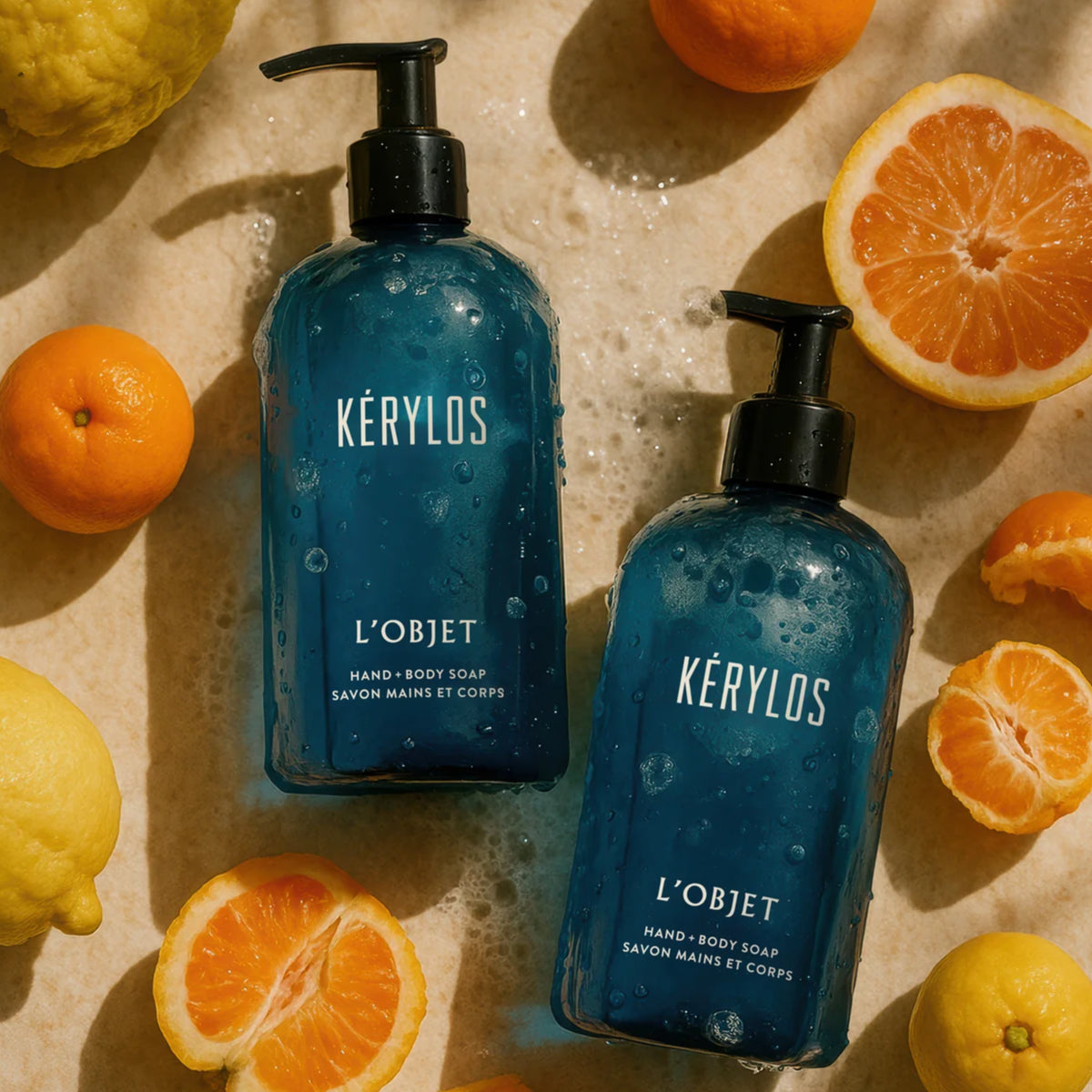 L'Objet - Kérylos - Hand & Body Soap in blauem Spender mit Wasser, umgeben von Zitrusfrüchten auf hellem Hintergrund