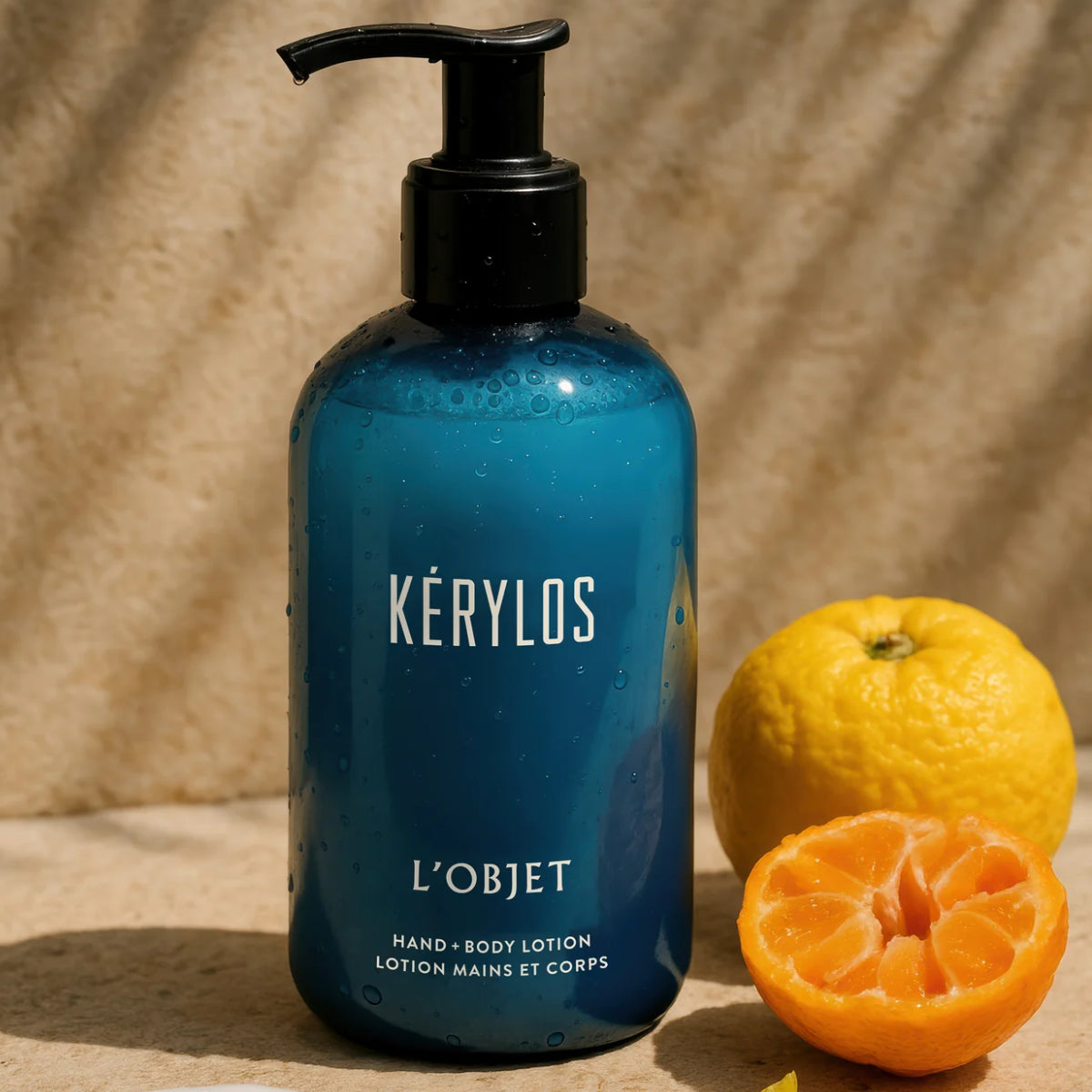 L'Objet - Kérylos - Hand & Body Lotion in blauem Spender mit Yuzu-Früchten auf hellem Hintergrund