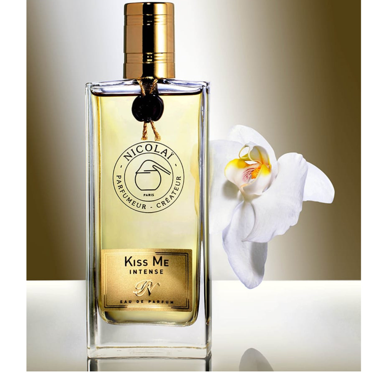 Nicolaï - Kiss Me Intense Parfümflakon mit goldenem Verschluss neben weißer Orchidee vor hellem Hintergrund