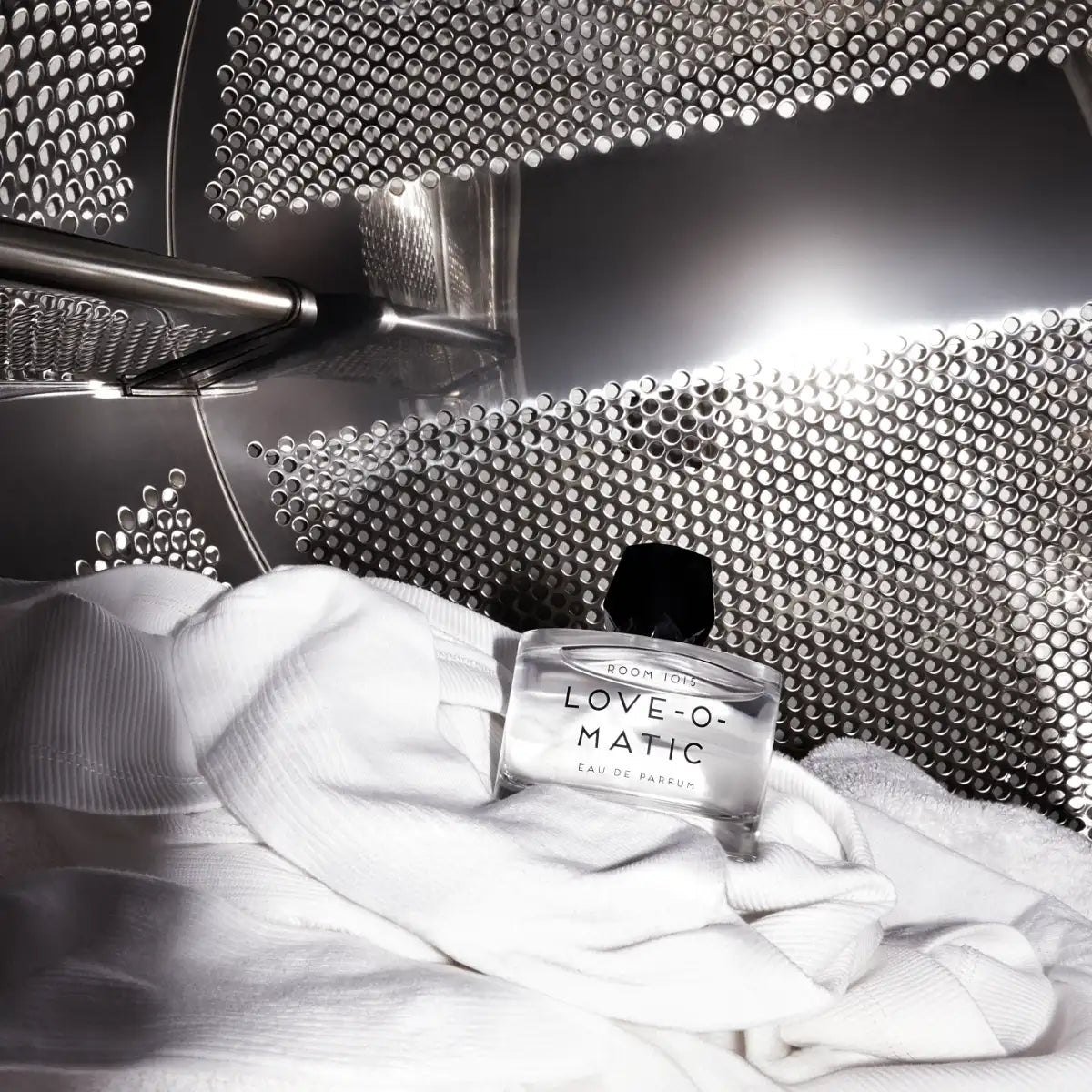 Room 1015 - Love-O-Matic Flakon auf weißem Textil in industriellem Waschmaschinentrommel-Setting kunstvoll präsentiert