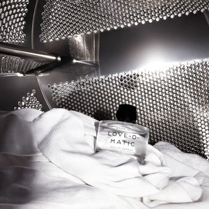 Room 1015 - Love-O-Matic Flakon auf weißem Textil in industriellem Waschmaschinentrommel-Setting kunstvoll präsentiert