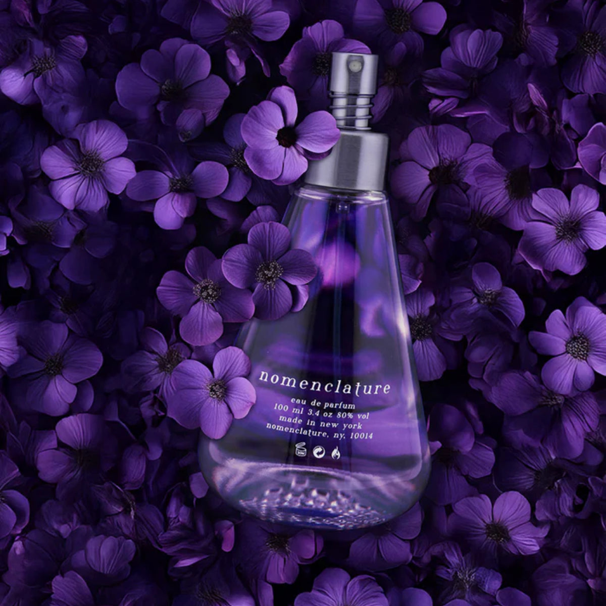 Nomenclature - lumen_esce Parfumflakon in violett mit silbernem Zerstäuber, umgeben von violetten Blütenblättern
