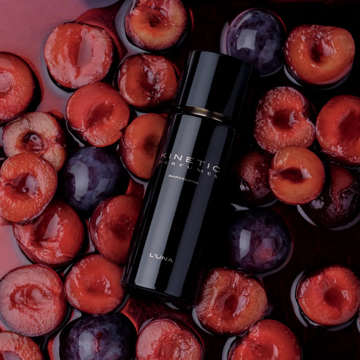 Kinetic Perfumes - L'Una schwarzer Flakon auf liegendem Bett aus halbierten Pflaumen mit glänzender Oberfläche