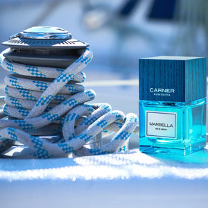 Carner Barcelona - Dream Collection - Marbella Parfumflakon in leuchtendem Blau mit holzstrukturierter Kappe vor maritimem Hintergrund