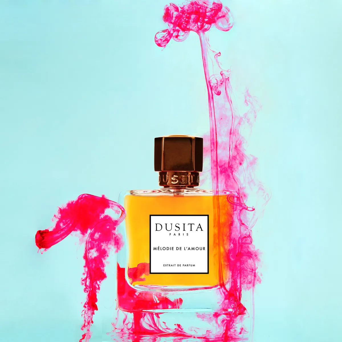 Dusita - Mélodie de l’Amour Parfumflakon mit bernsteinfarbener Essenz umgeben von rosa Tinte vor hellblauem Hintergrund