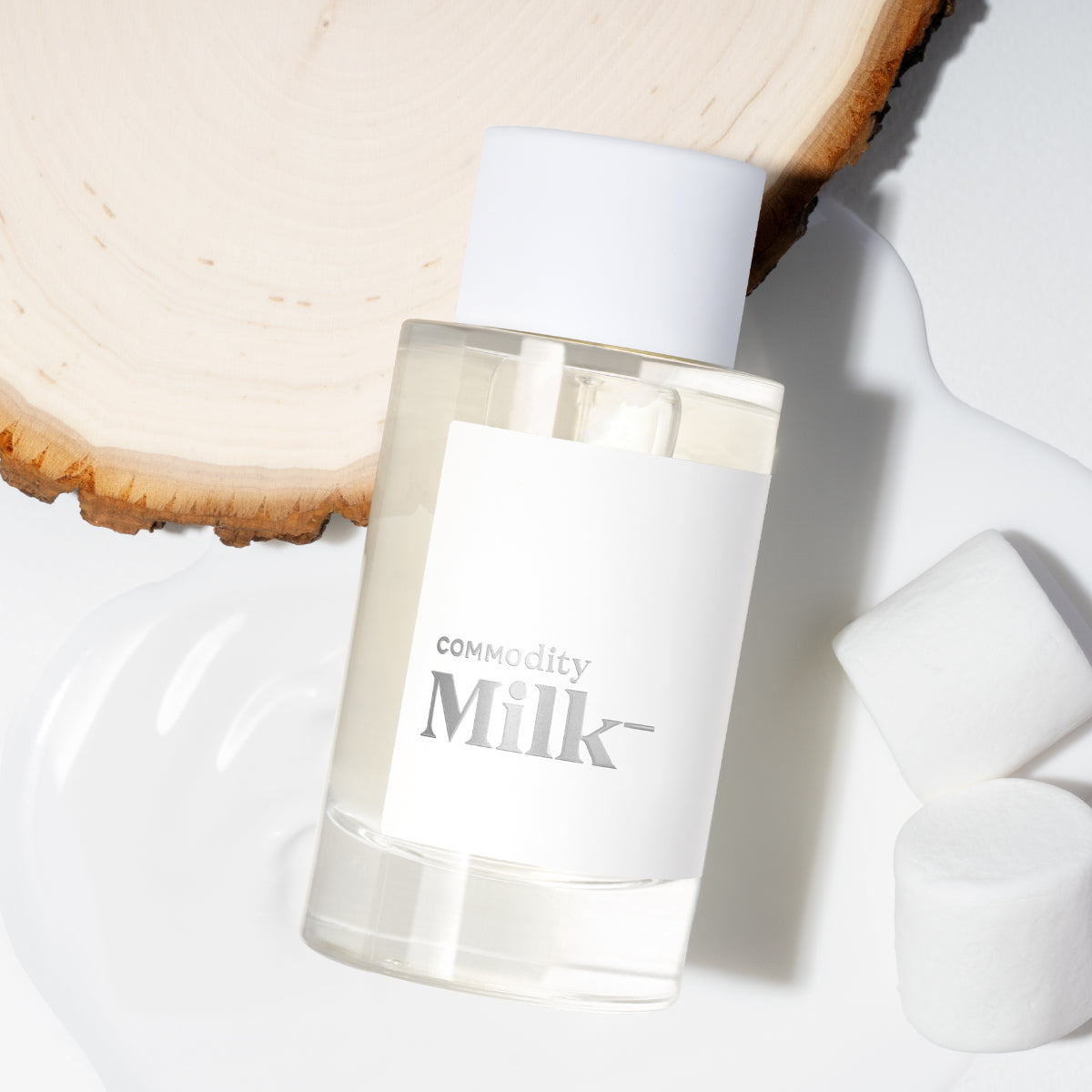 Commodity - Milk- Duftflakon mit minimalistischem weißen Etikett neben Holzscheibe und Marshmallow vor hellem Hintergrund