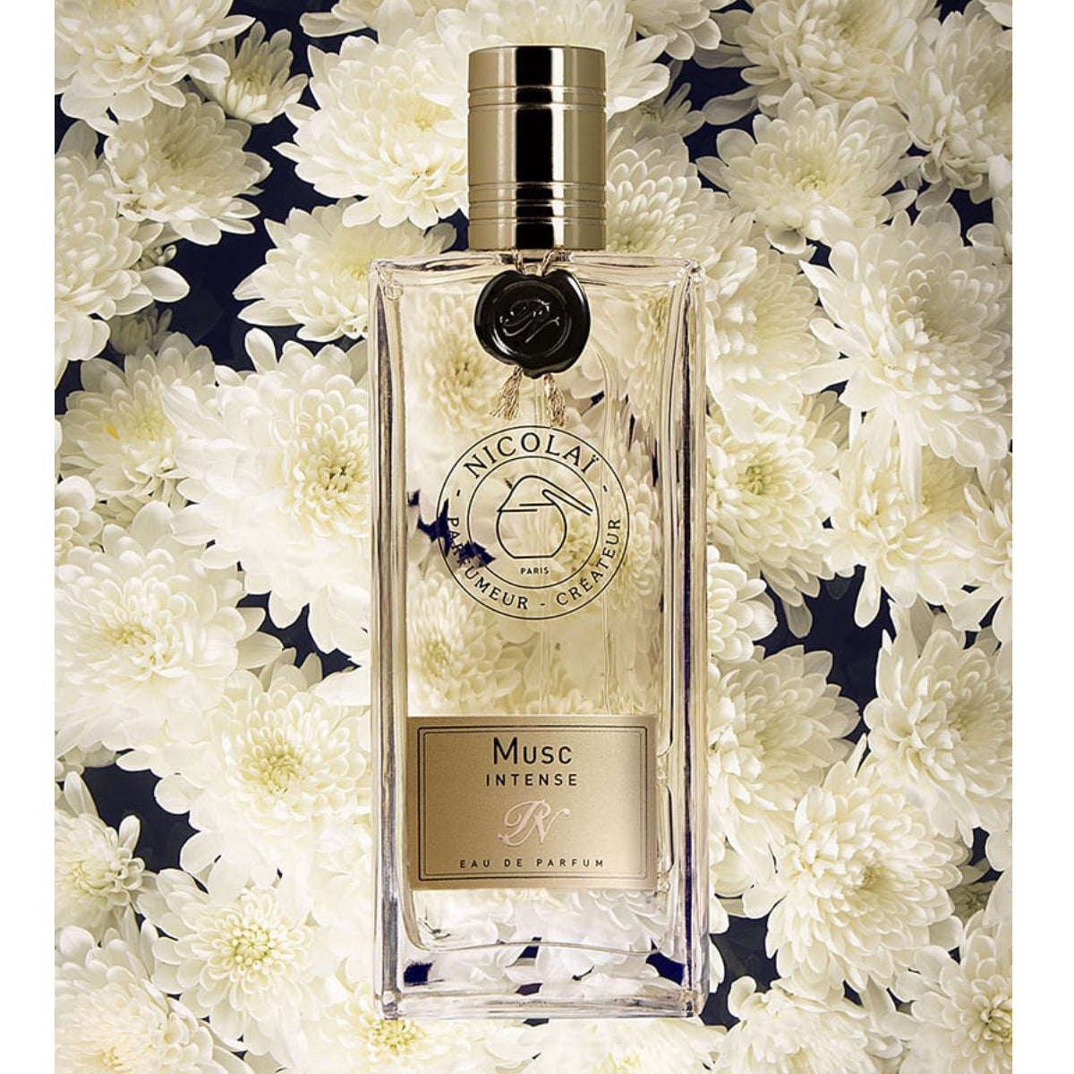 Nicolaï - Musc Intense Flakon mit goldenem Verschluss vor zarten weißen Blumen in elegantem Design
