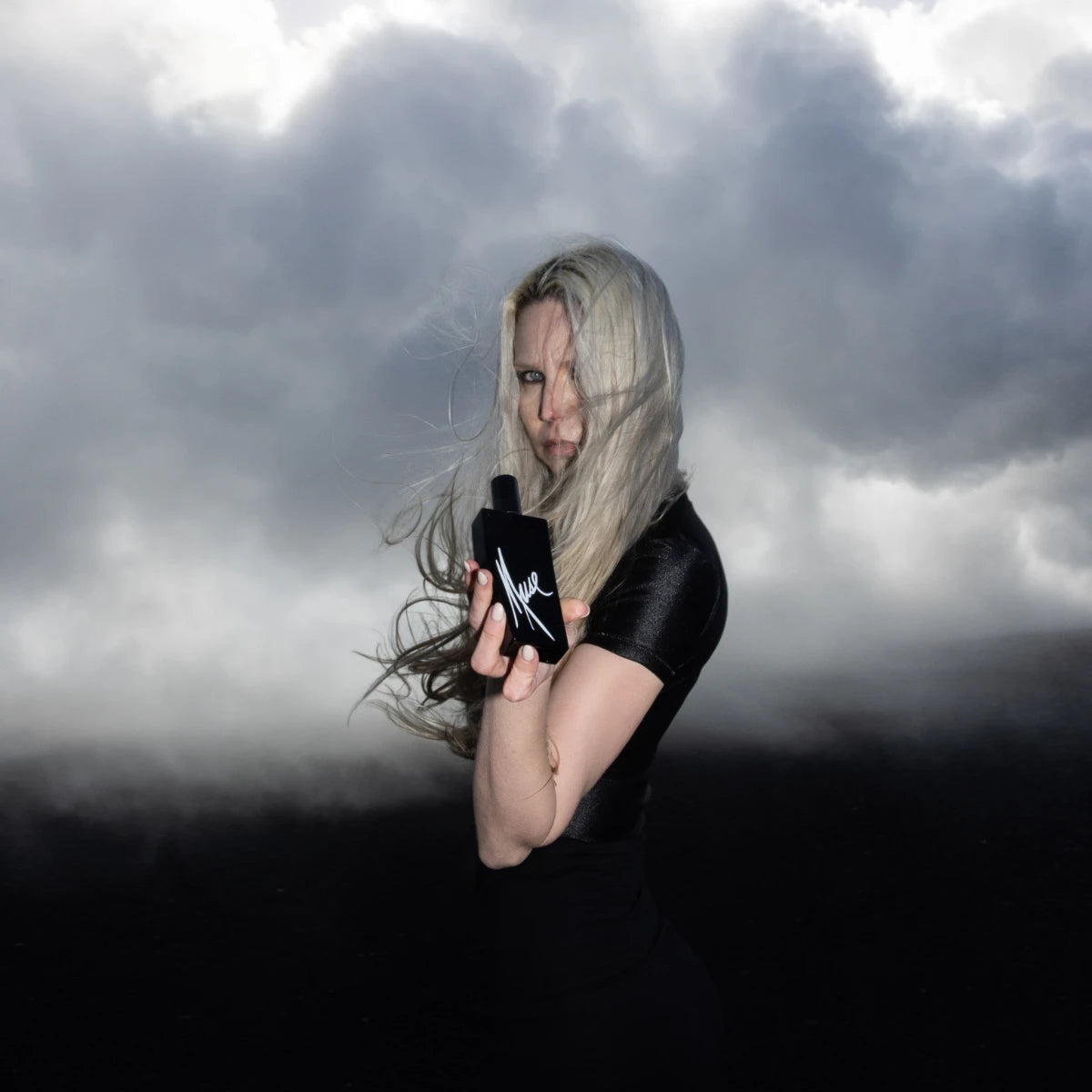 Andrea Maack Parfums - Muse in schwarzem Flakon gehalten von Frau vor dramatisch bewölktem Himmel