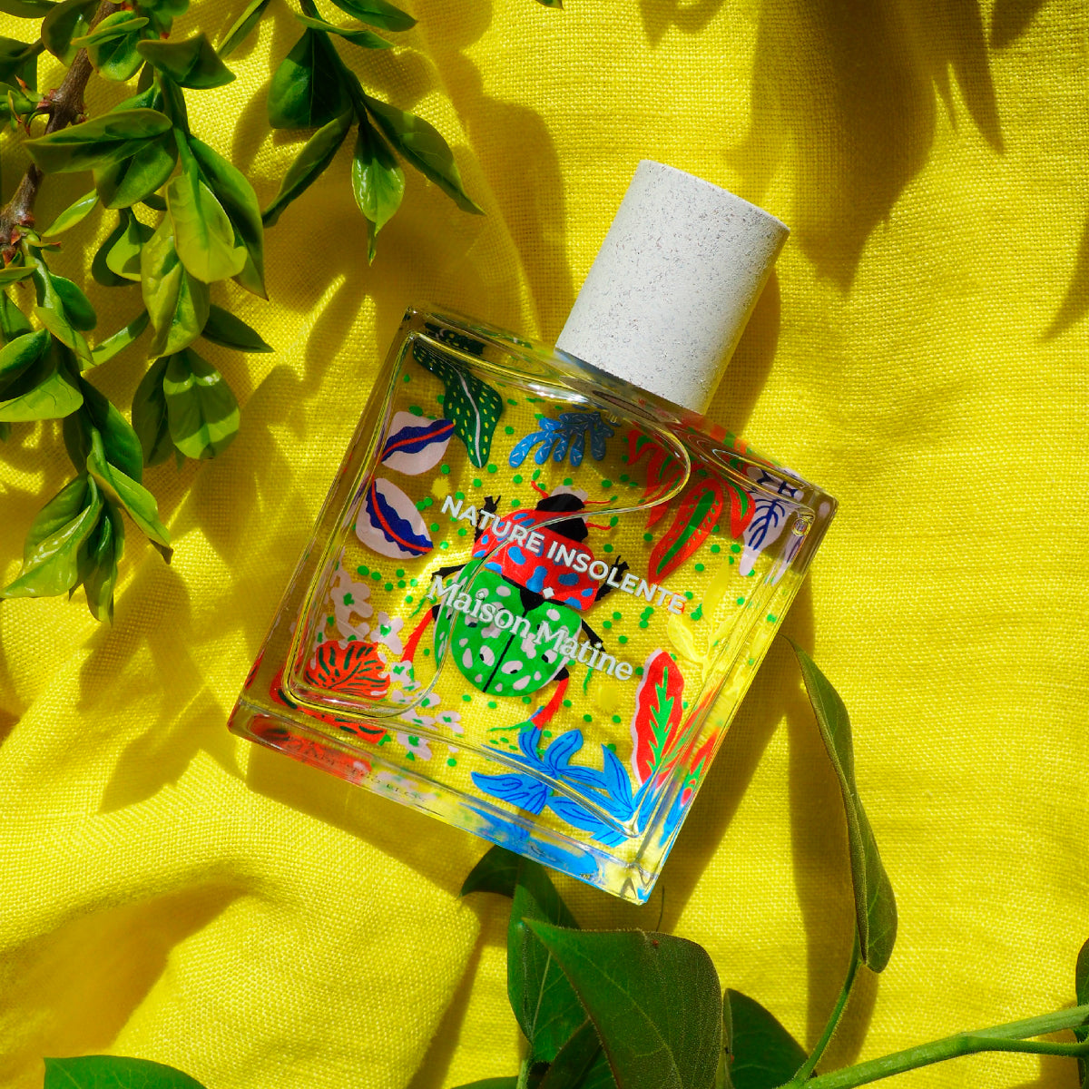 Maison Matine - Nature Insolente Parfumflakon mit farbenfrohen Natur- und Käfermotiven auf klarem Glas vor gelbem Stoff