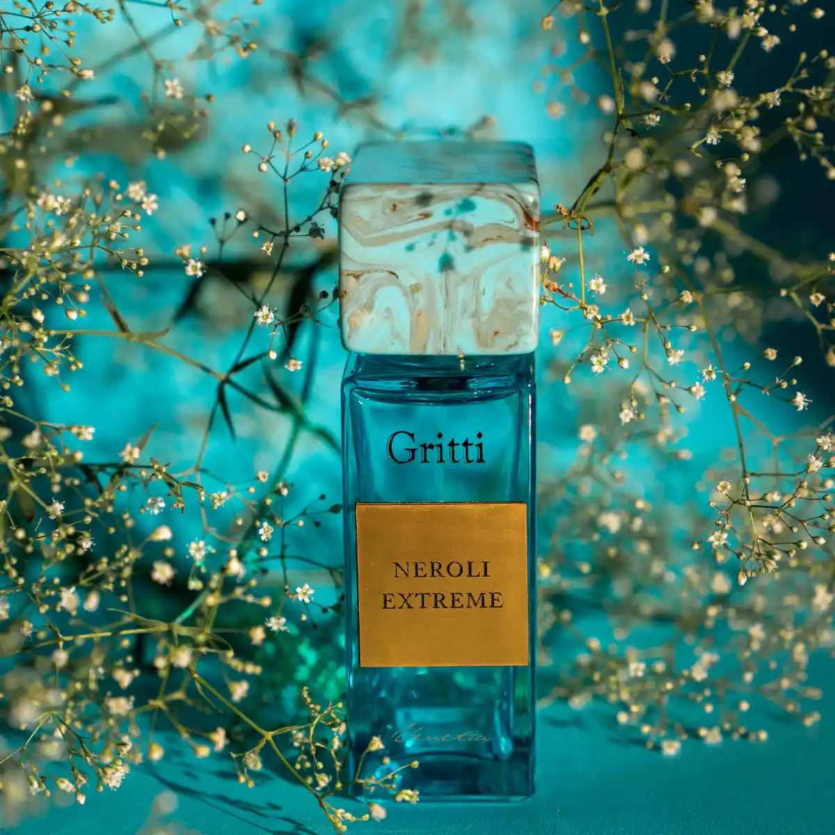 Gritti - Smaragd Collection - Neroli Extreme Parfumflakon in türkisblauem Glas mit goldfarbenem Etikett und zarten Blüten im Hintergrund