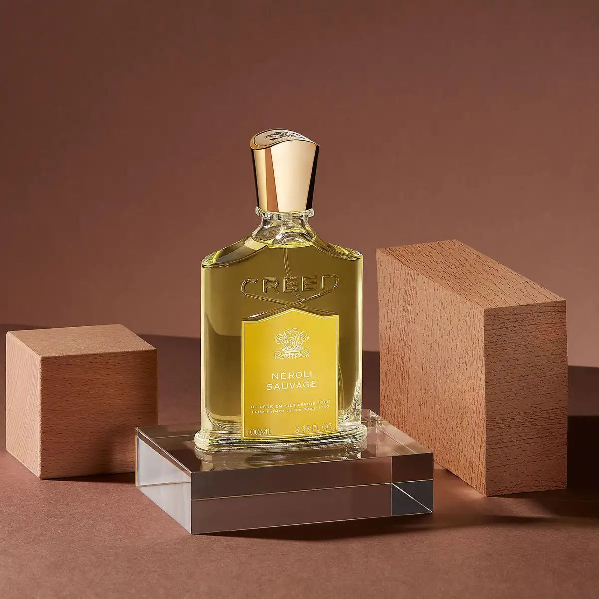 Creed - Neroli Sauvage Parfumflakon mit goldfarbenem Verschluss auf transparentem Podest und braunem Hintergrund