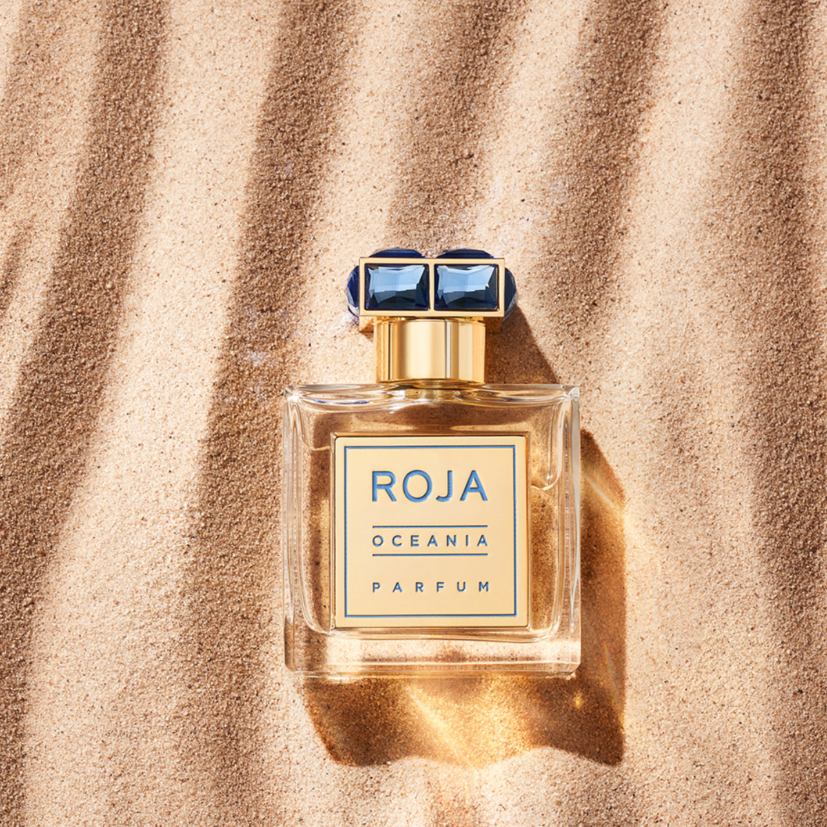 Roja Parfums - Oceania - Parfum Flakon mit blau-goldenem Etikett auf goldfarbenem Sand mit Sonnenlichtspiel