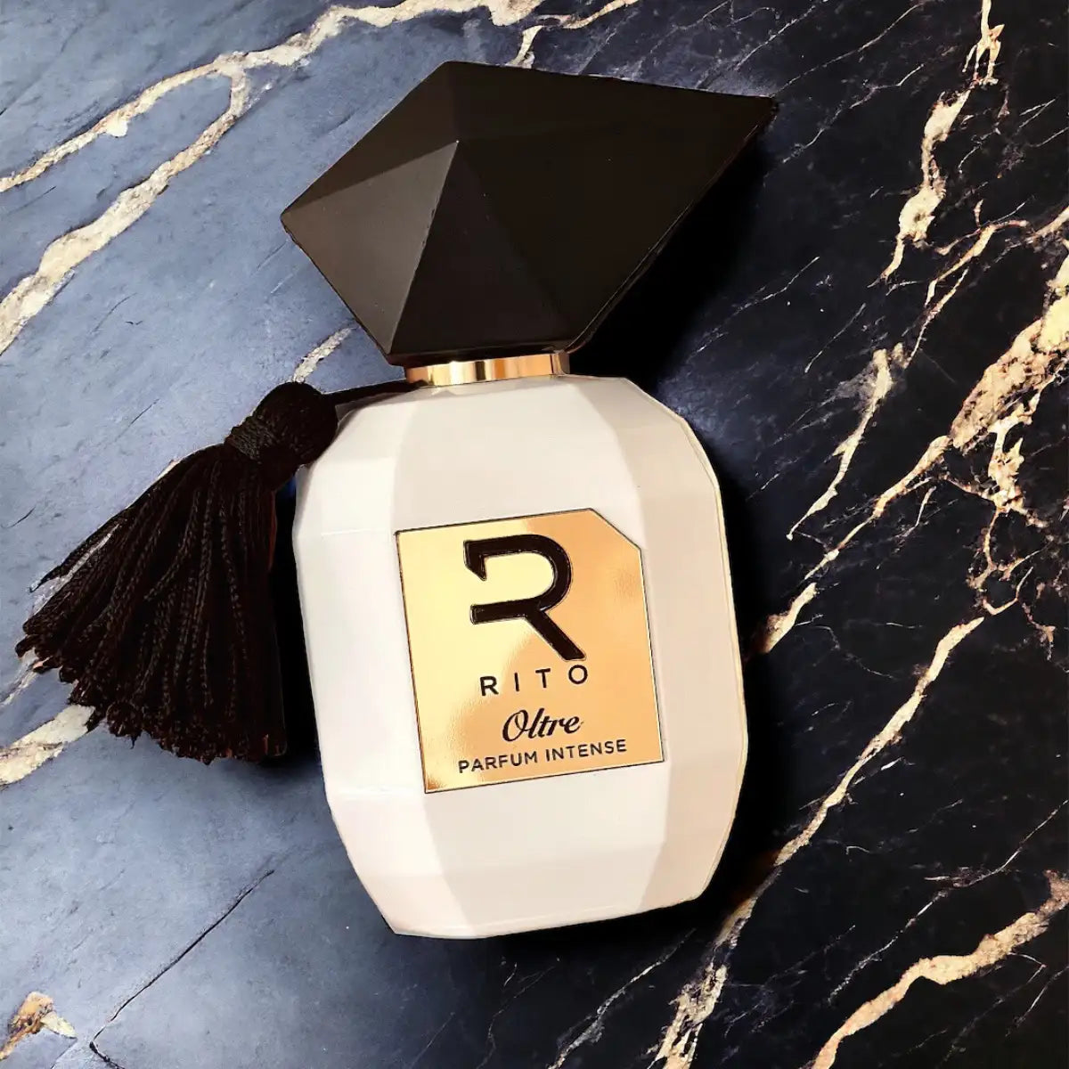 Rito - Oltre eleganter weißer Flakon mit facettiertem schwarzem Verschluss, goldfarbenem Label und dekorativer Quaste auf dunklem Marmor