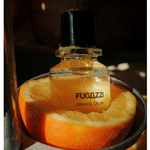 Fugazzi - Orange Crush Extrait Parfumflakon im Glas mit schwarzem Verschluss auf orangefarbener Frucht im Schattenlicht