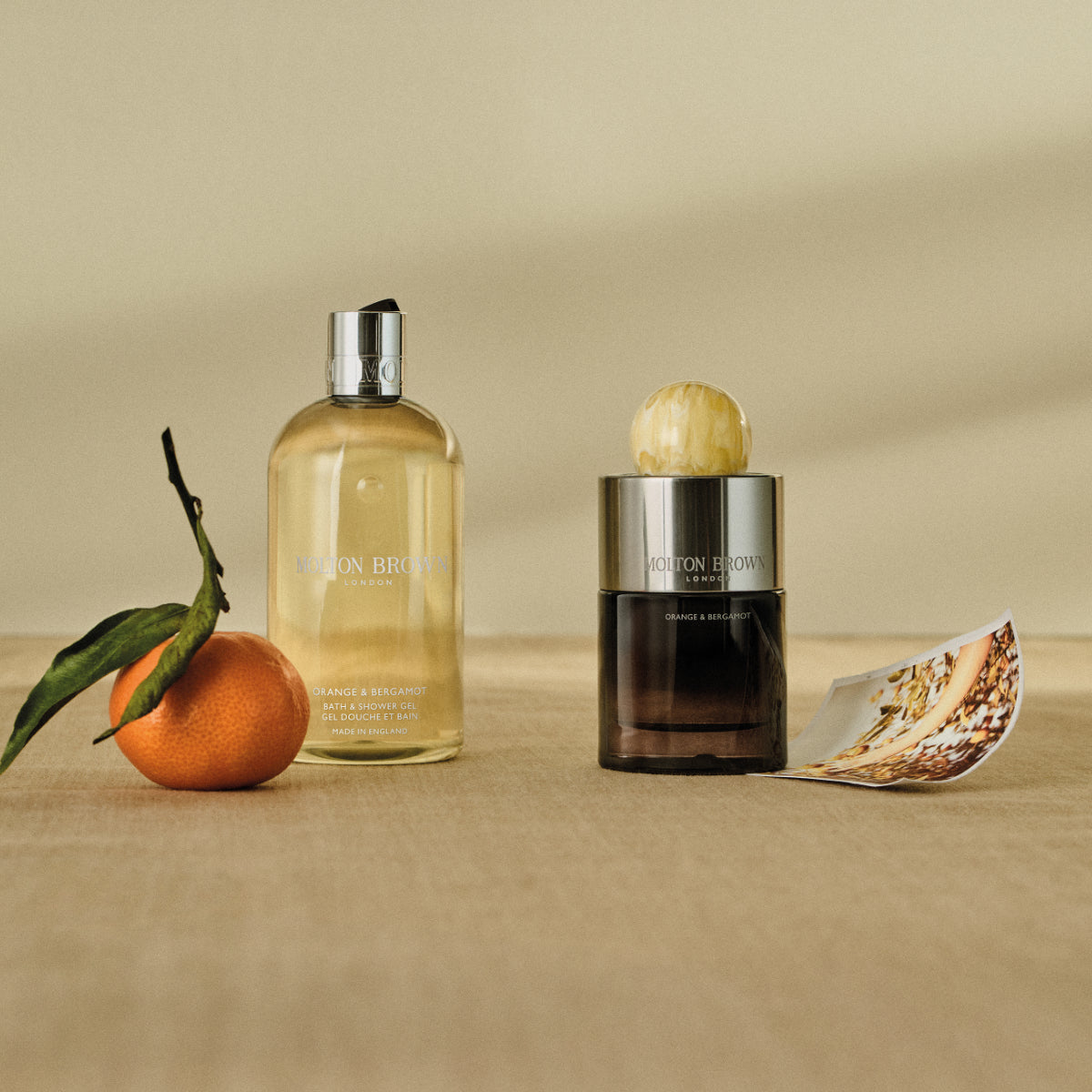 Molton Brown - Orange & Bergamot mit Duschgelflakon, Parfumflakon und frischer Mandarine vor neutralem Hintergrund