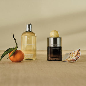 Molton Brown - Orange & Bergamot mit Duschgelflakon, Parfumflakon und frischer Mandarine vor neutralem Hintergrund