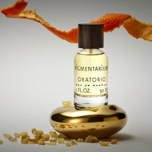 Pigmentarium - Oratorio Parfumflakon mit klarem Glas und dunklem Holzverschluss vor grauem Hintergrund mit Orangenschale und Harzperlen