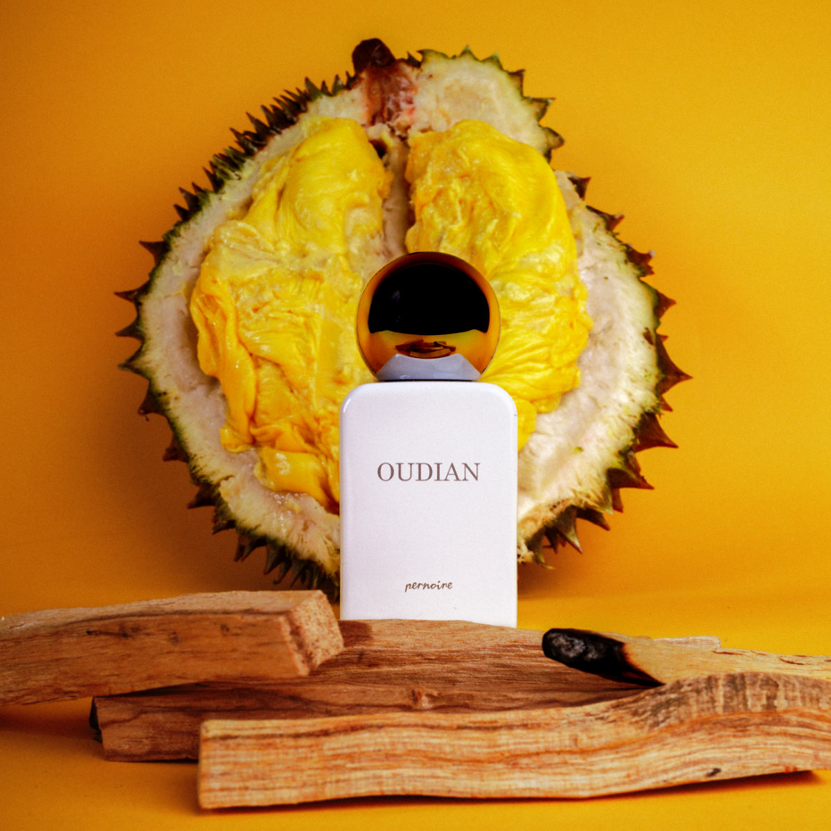 Pernoire - Odus Collection - Oudian Parfumflakon in weiß mit goldenem Verschluss vor exotischer Frucht und Holzdekoration