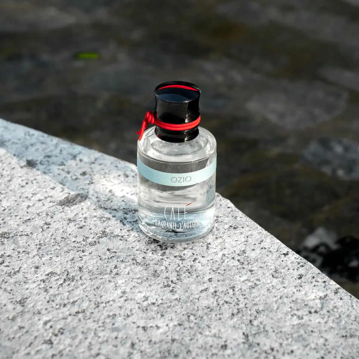 Calé Fragranze d'Autore - Ozio klarer Glasflakon mit schwarzem Verschluss und rotem Band auf hellem Stein unter Tageslicht