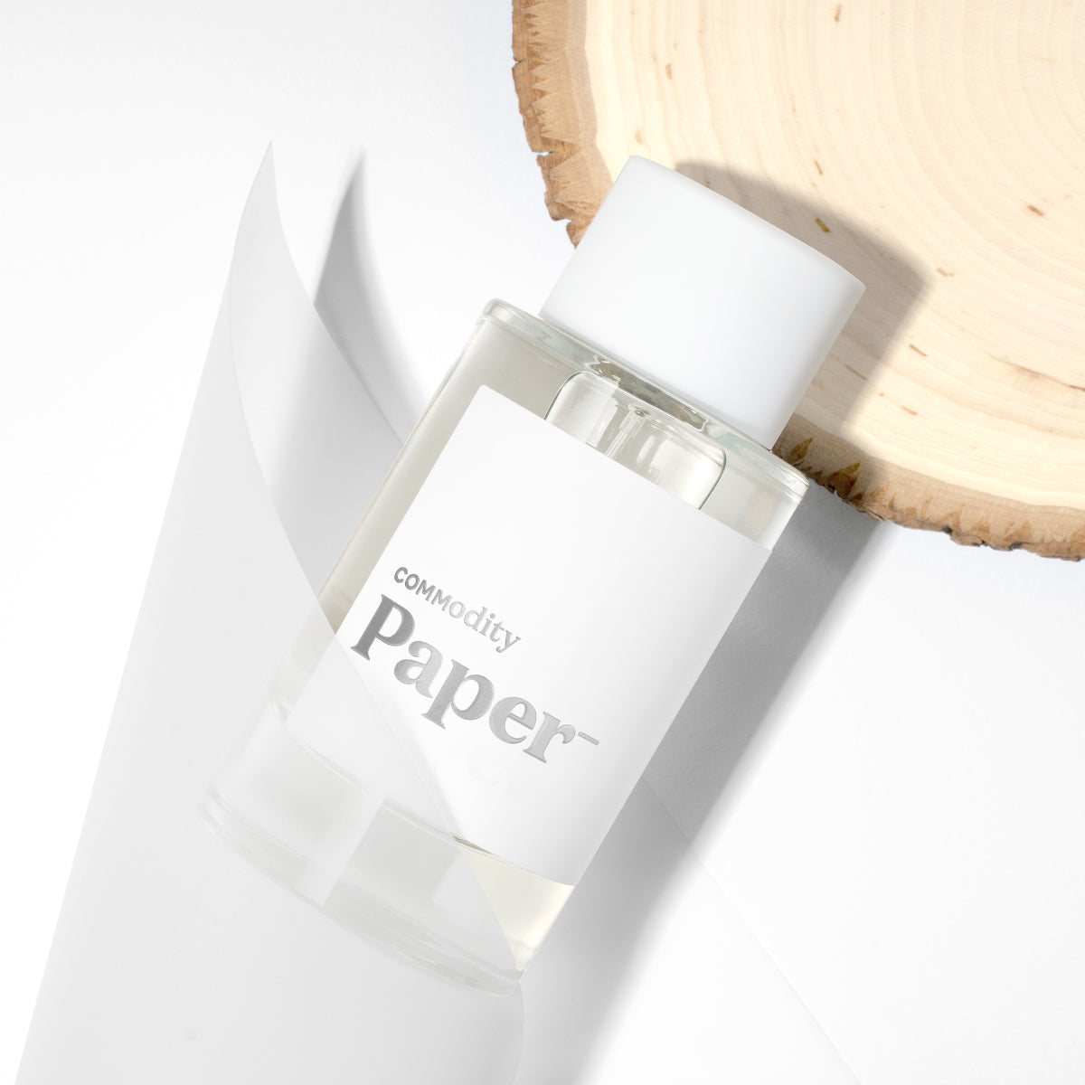 Commodity - Paper- Duftflakon mit minimalistischem weißen Etikett neben Holzscheibe auf hellem Hintergrund