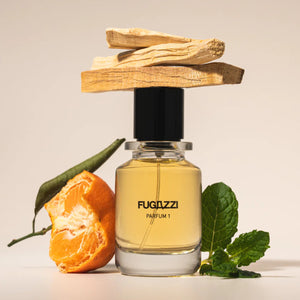 Fugazzi - Parfum 1 mit klarer Flasche, schwarzem Deckel, Mandarine, Holz und frischen grünen Blättern vor hellem Hintergrund