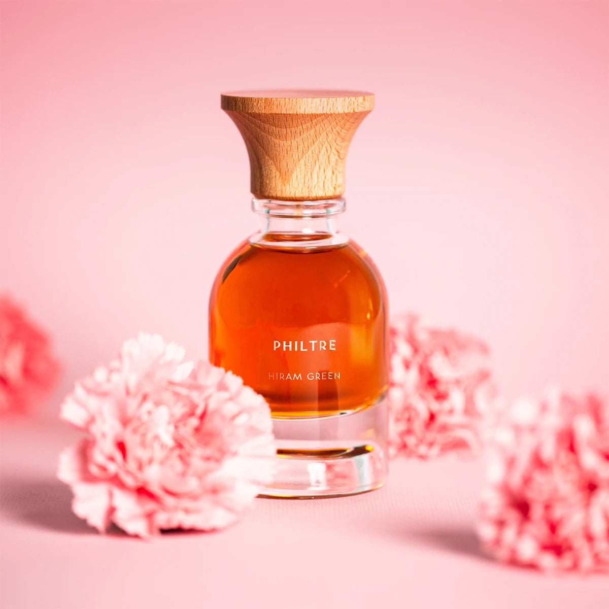 Hiram Green - Philtre Parfumflakon mit bernsteinfarbener Flüssigkeit und Holzverschluss vor zartrosa Blumendekor