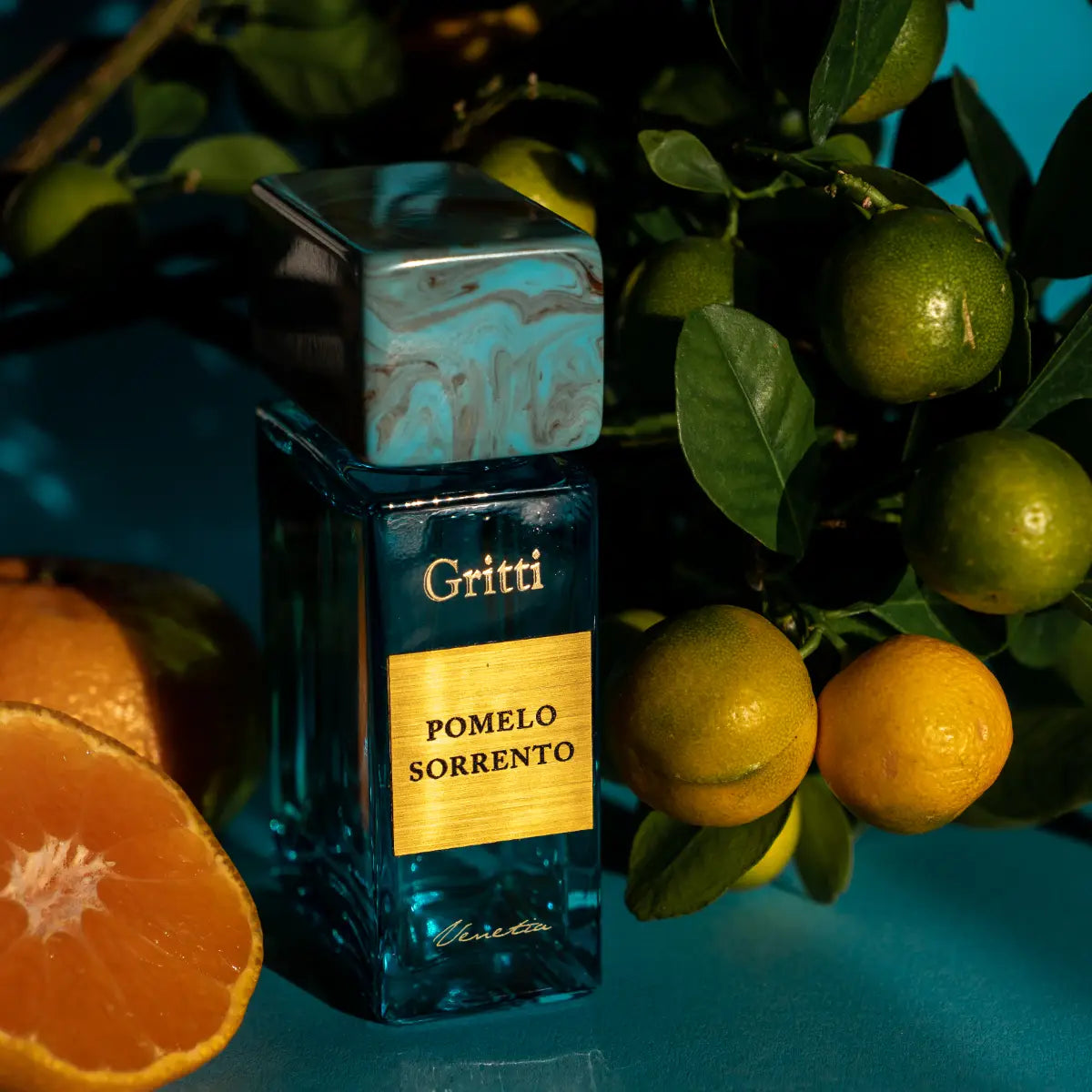 Gritti - Smaragd Collection - Pomelo Sorrento Parfumflakon in blaugrünem Glas mit goldfarbener Plakette und Zitrusfrüchten im Hintergrund