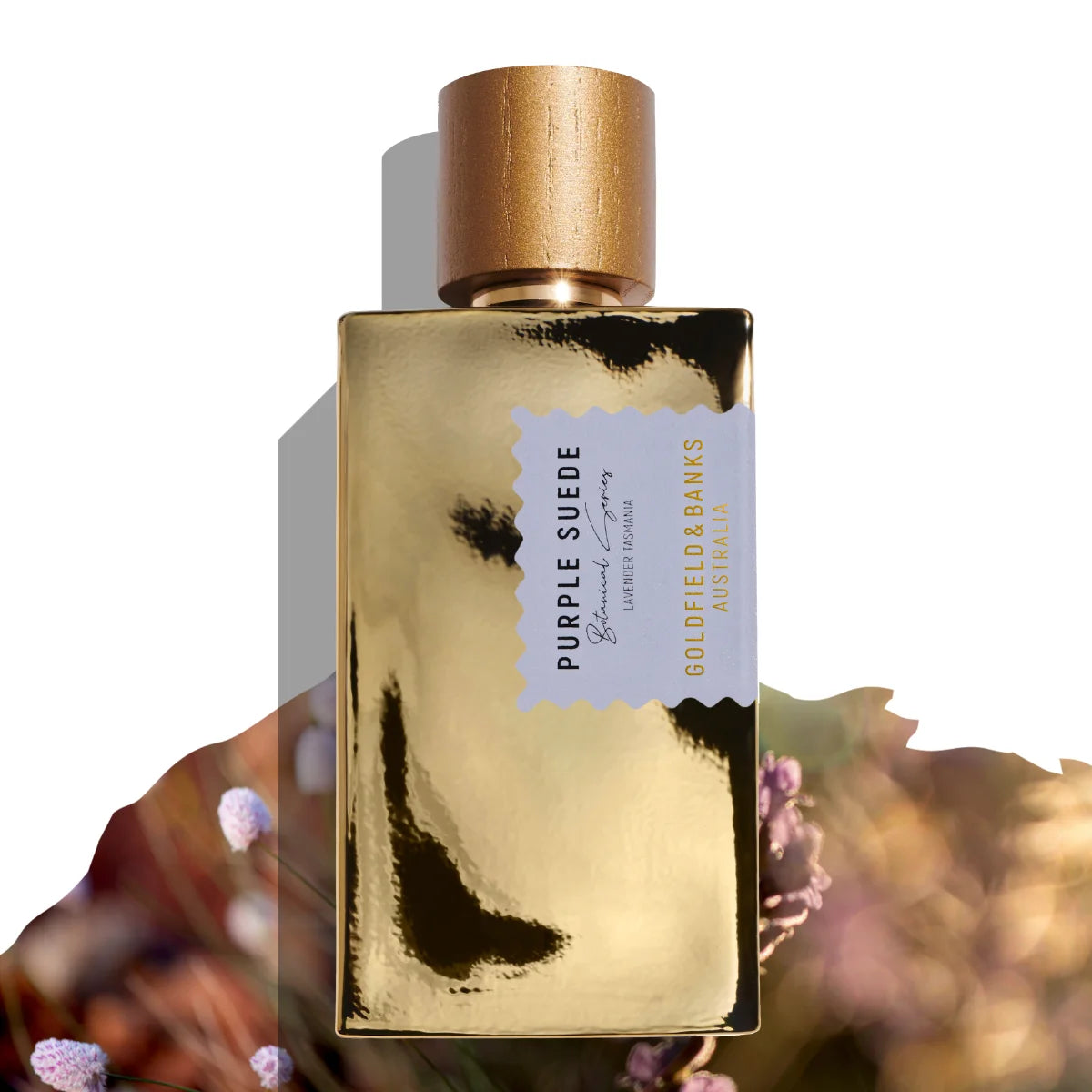 Goldfield & Banks - Native Collection - Purple Suede Parfumflakon mit goldener Oberfläche und hölzernem Verschluss vor floralem Hintergrund