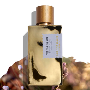 Goldfield & Banks - Native Collection - Purple Suede Parfumflakon mit goldener Oberfläche und hölzernem Verschluss vor floralem Hintergrund
