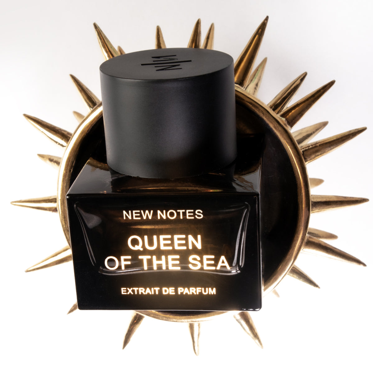 New Notes - Contemporary Blend Collection - Queen of the Sea schwarzer Parfumflakon vor dekorativer goldener Sonnenform