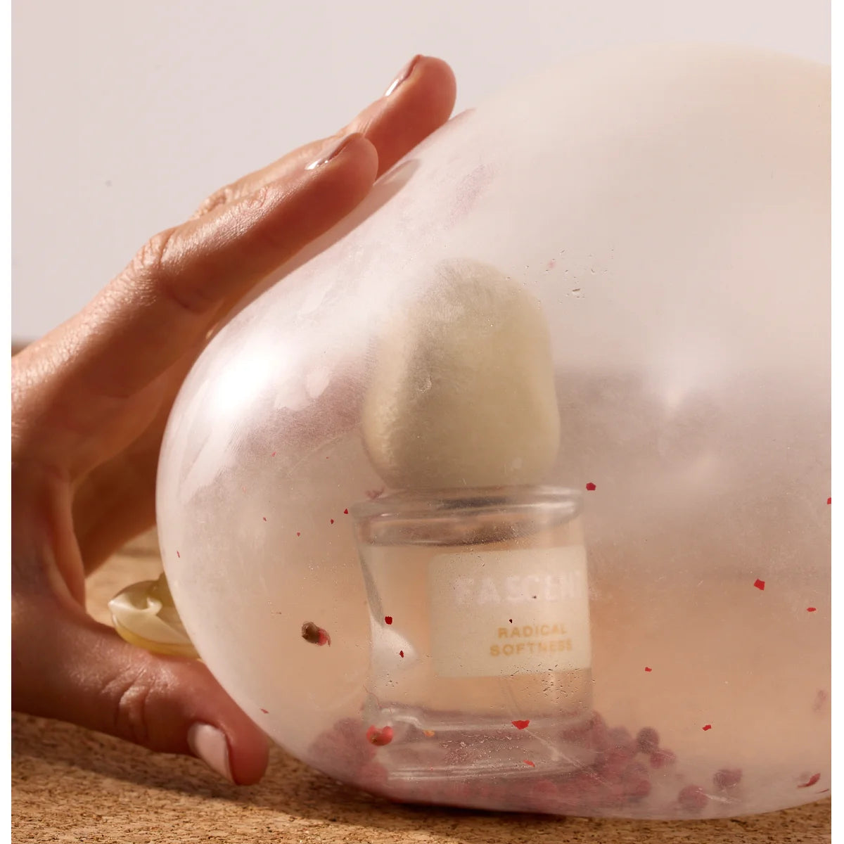 Fascent - Radical Softness Parfumflakon mit samtigem Deckel im transparenten Ballon mit roten Akzenten und zarter Hand davor