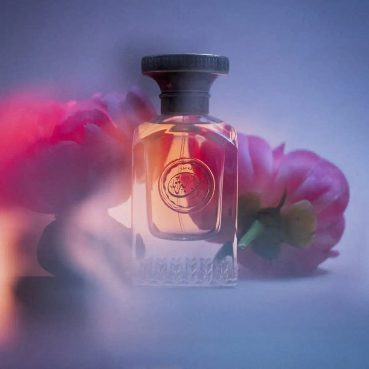 Anfas - Essence of Arabia Collection - Rahaba Parfumflakon mit goldenem Inhalt vor zart unscharfem Hintergrund und Blumenarrangement
