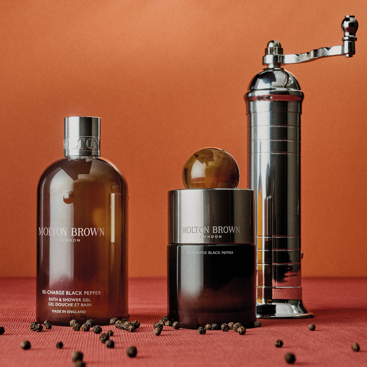 Molton Brown - Re-Charge Black Pepper Set mit Duschgel, Duftflakon mit rundem Verschluss und silberner Pfeffermühle vor orangefarbenem Hintergrund