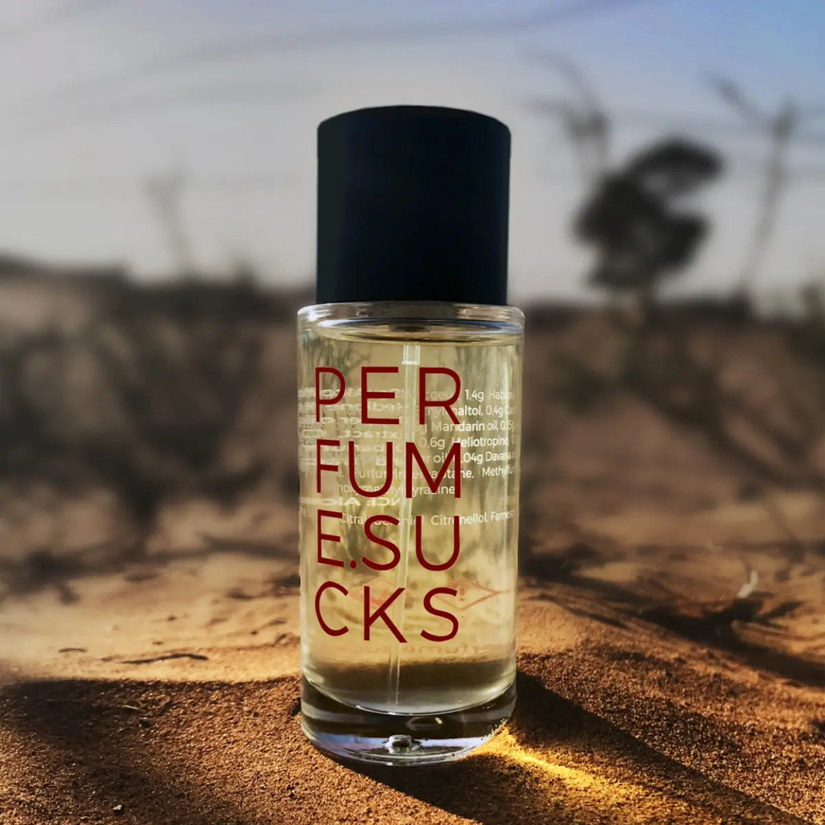 PERFUME.SUCKS - Red 198C Parfumflakon mit schwarzem Verschluss und roter Schrift auf sandigem Untergrund bei Sonnenuntergang