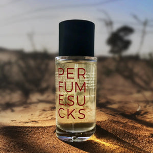 PERFUME.SUCKS - Red 198C Parfumflakon mit schwarzem Verschluss und roter Schrift auf sandigem Untergrund bei Sonnenuntergang