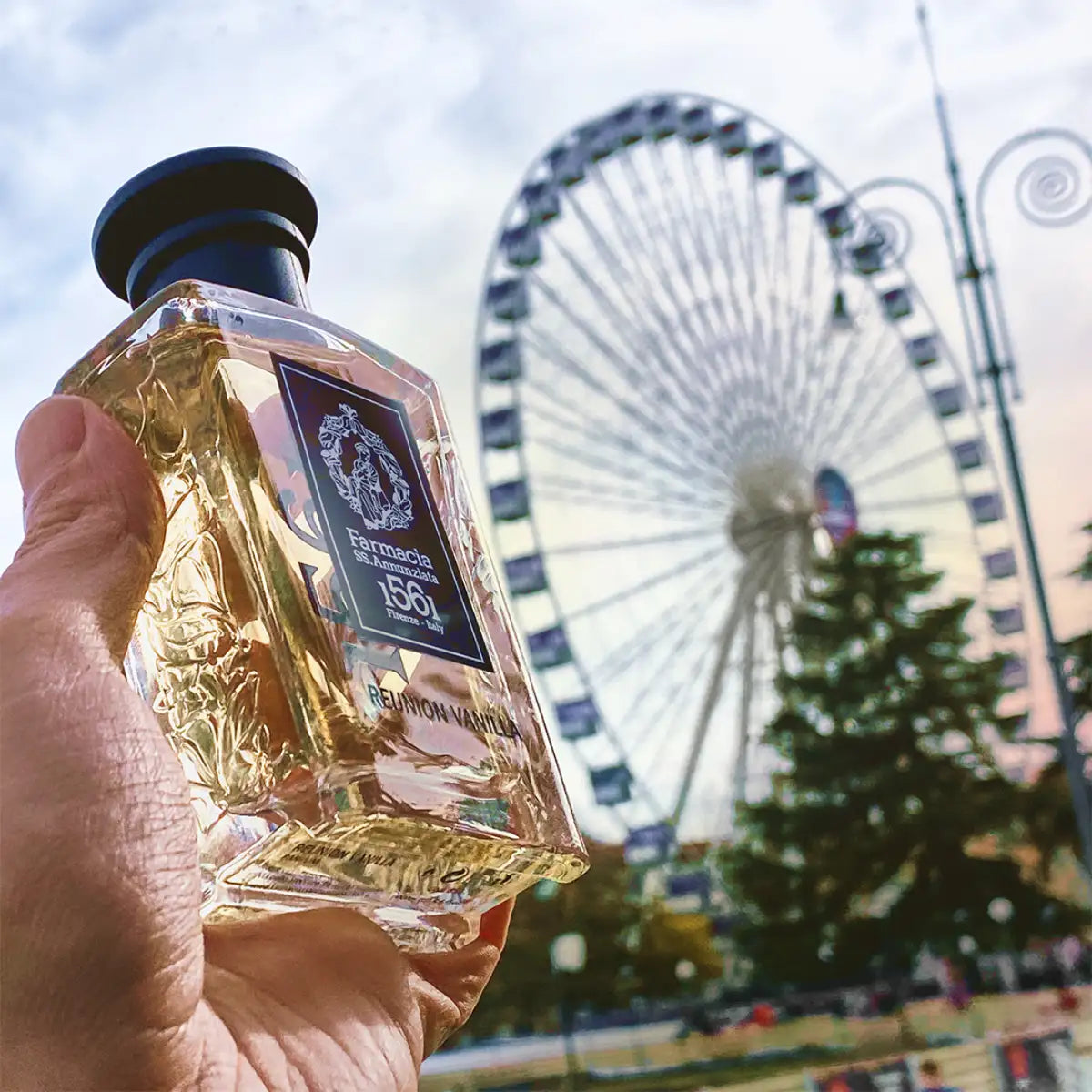 Farmacia SS. Annunziata - Reunion Vanilla Parfumflakon mit goldenem Duft vor Riesenrad und Himmel im Hintergrund