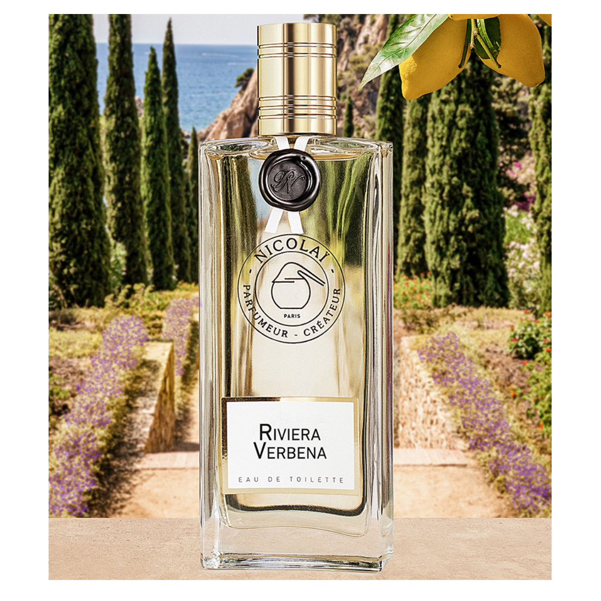 Nicolaï - Riviera Verbena klarer Flakon mit goldenem Verschluss vor mediterraner Gartenkulisse und Zitrusfrüchten
