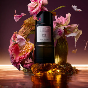 D'Orsay - Rose Blaze Extrait de Parfum in schwarzem Flakon umgeben von rosa Blüten und warmem Licht vor dunklem Hintergrund
