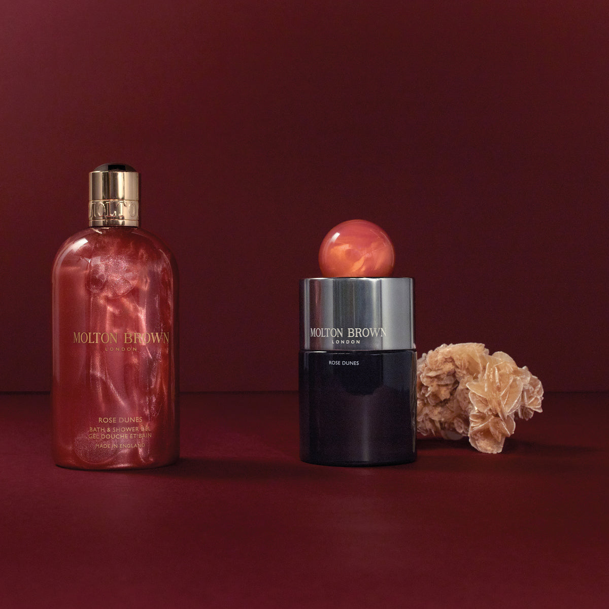 Molton Brown - Rose Dunes Duschgel und eleganter Duftzerstäuber vor tiefrotem Hintergrund mit natürlichem Dekor