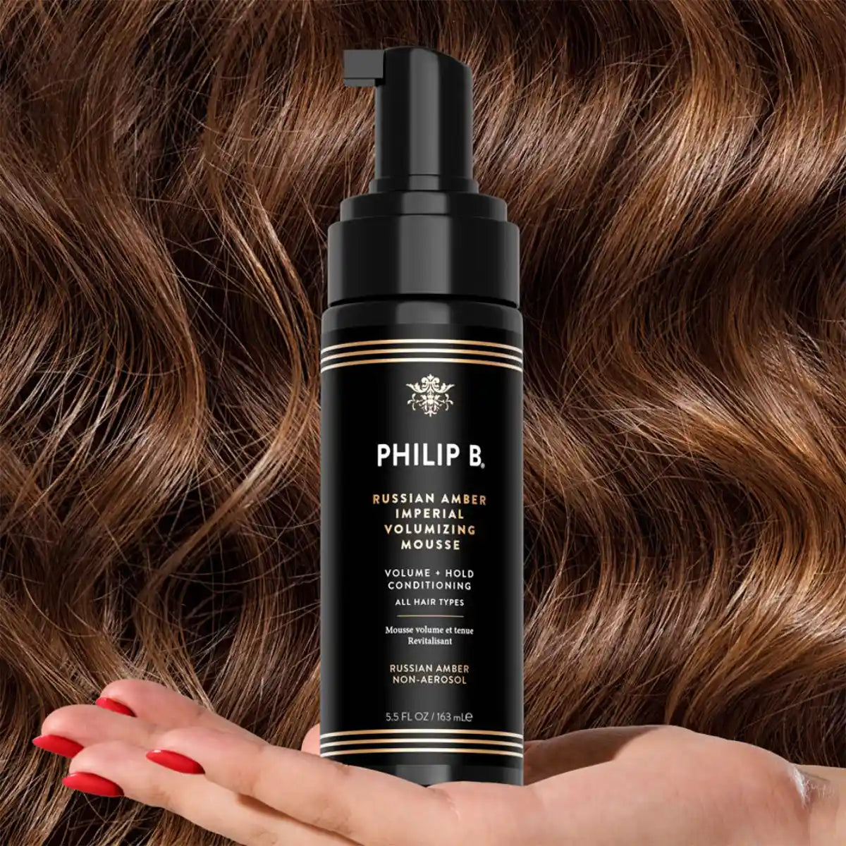 Philip B - Russian Amber Imperial Volumizing Mousse Non-Aerosol in schwarzer Flasche vor welligem braunem Haar mit rotem Nagellack